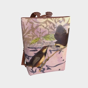 Puede incluir: Una mochila rosa y marrón con un estampado floral y dos pájaros posados en una rama. Los pájaros son marrones y negros con detalles amarillos.
