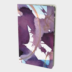 Op de afbeelding: Een hardcover notitieboek met een aquarelontwerp. De omslag toont abstracte paarse, gouden en blauwe penseelstreken op een witte achtergrond. Het notitieboek is rechthoekig en heeft een strakke, moderne uitstraling.