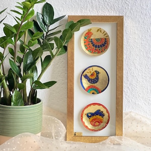 Pode incluir: Uma obra de arte emoldurada com tr&ecirc;s pratos decorativos circulares dourados. Cada prato apresenta um desenho de p&aacute;ssaro colorido com detalhes em vermelho, azul e amarelo. A moldura &eacute; marrom claro e o fundo &eacute; branco.