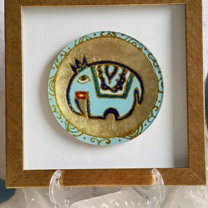 Pode incluir: Obra de arte emoldurada com um design de elefante azul claro e detalhes dourados. O elefante tem uma coroa e padr&otilde;es decorativos. A obra de arte est&aacute; sobre um fundo dourado e emoldurada em uma moldura de madeira dourada.