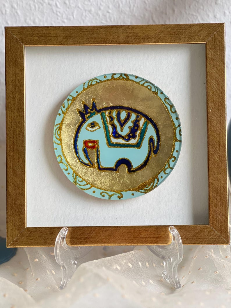 Pode incluir: Obra de arte emoldurada com um design de elefante azul claro e detalhes dourados. O elefante tem uma coroa e padr&otilde;es decorativos. A obra de arte est&aacute; sobre um fundo dourado e emoldurada em uma moldura de madeira dourada.