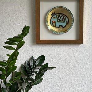 Pode incluir: Um prato decorativo redondo emoldurado com um desenho de elefante azul claro e detalhes dourados. O elefante tem uma coroa e padr&otilde;es decorativos. O prato est&aacute; sobre um fundo branco dentro de uma moldura de madeira, montado em uma parede branca texturizada.