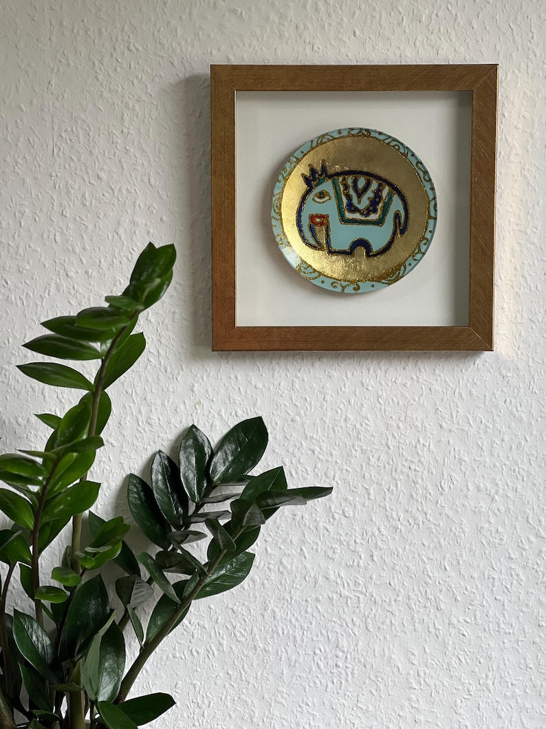Pode incluir: Um prato decorativo redondo emoldurado com um desenho de elefante azul claro e detalhes dourados. O elefante tem uma coroa e padr&otilde;es decorativos. O prato est&aacute; sobre um fundo branco dentro de uma moldura de madeira, montado em uma parede branca texturizada.