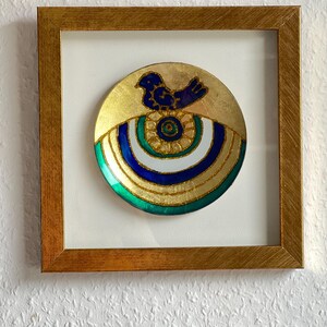 Pode incluir: Prato decorativo emoldurado com fundo dourado e desenho de p&aacute;ssaro azul. O prato apresenta an&eacute;is conc&ecirc;ntricos verdes, brancos e azuis, e est&aacute; envolto em uma moldura de madeira dourada. A obra de arte &eacute; exibida em um fundo branco.