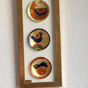 Pode incluir: Uma obra de arte emoldurada com tr&ecirc;s pequenos pratos decorativos redondos. Cada prato tem um fundo dourado com desenhos coloridos de p&aacute;ssaros em vermelho, azul, laranja e verde. A moldura &eacute; de cor dourada quente.