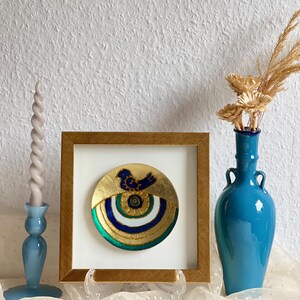 Pode incluir: Uma pe&ccedil;a decorativa redonda emoldurada, com fundo dourado e um desenho de p&aacute;ssaro azul. A pe&ccedil;a &eacute; emoldurada em uma moldura dourada e exibida em um suporte transparente. Um vaso azul com flores secas e um casti&ccedil;al azul tamb&eacute;m est&atilde;o presentes.