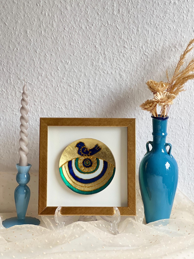 Pode incluir: Uma pe&ccedil;a decorativa redonda emoldurada, com fundo dourado e um desenho de p&aacute;ssaro azul. A pe&ccedil;a &eacute; emoldurada em uma moldura dourada e exibida em um suporte transparente. Um vaso azul com flores secas e um casti&ccedil;al azul tamb&eacute;m est&atilde;o presentes.