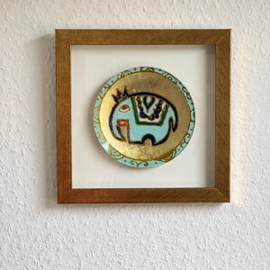 Pode incluir: Um prato decorativo circular emoldurado com um design de elefante azul claro com detalhes dourados e azul marinho. O prato est&aacute; sobre um fundo branco dentro de uma moldura de madeira, criando uma pe&ccedil;a de arte de parede visualmente atraente.