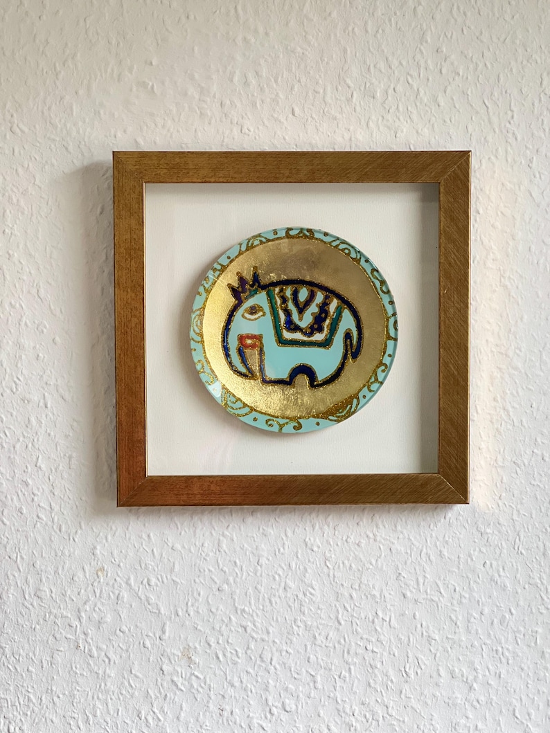 Pode incluir: Um prato decorativo circular emoldurado com um design de elefante azul claro com detalhes dourados e azul marinho. O prato est&aacute; sobre um fundo branco dentro de uma moldura de madeira, criando uma pe&ccedil;a de arte de parede visualmente atraente.