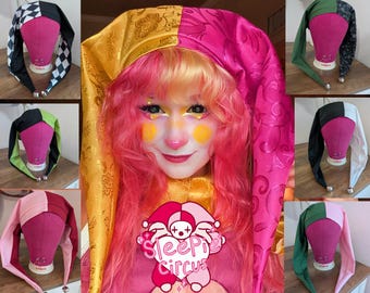 May include: A collection of colorful jester hats in various color combinations, including black and white checkered, pink and gold, and green and black. Each hat has long, pointed ends with a small silver bell at the tip. The hats are displayed on pink mannequin heads.