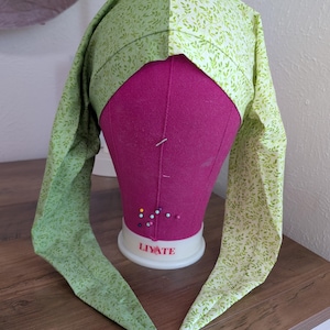 May include: A green and white patterned bonnet with long ties, displayed on a pink mannequin head. The fabric features a delicate leaf design. The bonnet is likely handmade and designed for comfort and style.