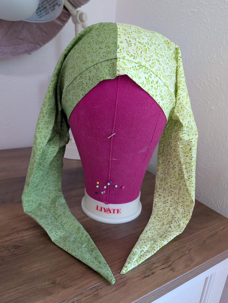 May include: A green and white patterned bonnet with long ties, displayed on a pink mannequin head. The fabric features a delicate leaf design. The bonnet is likely handmade and designed for comfort and style.