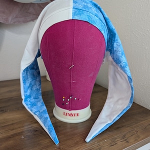 May include: A white and blue bonnet with long, pointed ear-like flaps, displayed on a pink mannequin head. The bonnet is divided into two sections: one white and the other with a blue floral pattern. The mannequin is on a wooden surface.