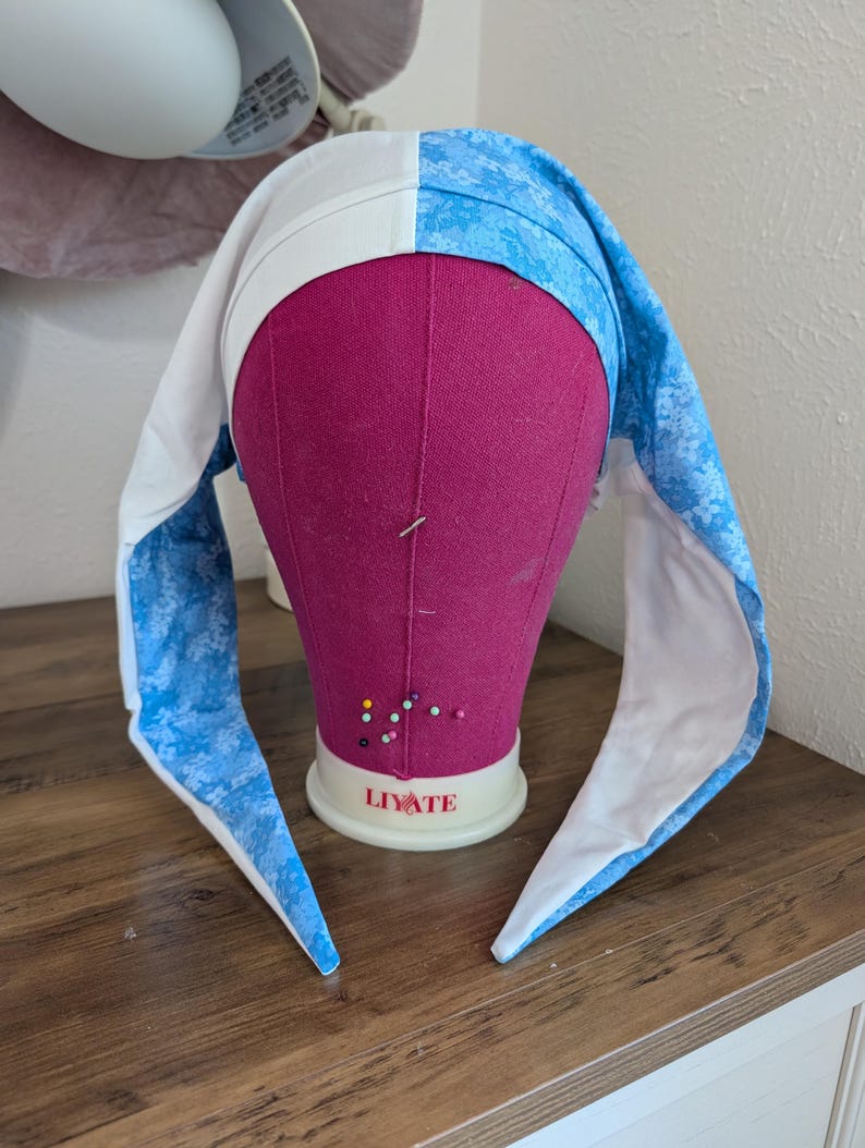 May include: A white and blue bonnet with long, pointed ear-like flaps, displayed on a pink mannequin head. The bonnet is divided into two sections: one white and the other with a blue floral pattern. The mannequin is on a wooden surface.