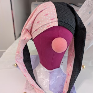 May include: A jester's hat with a pink and black color scheme, featuring a polka dot pattern with bat silhouettes. The hat is displayed on a purple mannequin head with a pink nose. The collar is a patchwork of white and lavender fabric.