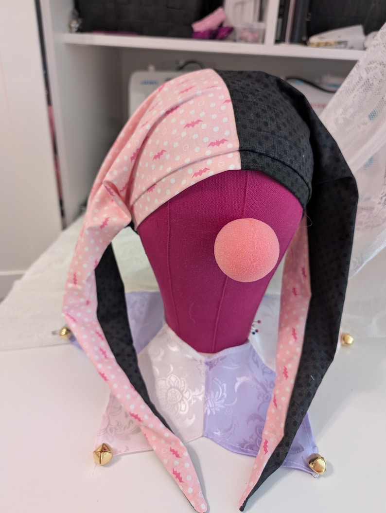 May include: A jester's hat with a pink and black color scheme, featuring a polka dot pattern with bat silhouettes. The hat is displayed on a purple mannequin head with a pink nose. The collar is a patchwork of white and lavender fabric.