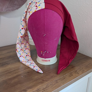 May include: A handmade jester hat with a two-tone design. One side features a patchwork of colorful teardrop shapes on a light pink background, while the other side is a solid deep red. The hat is displayed on a pink mannequin head.