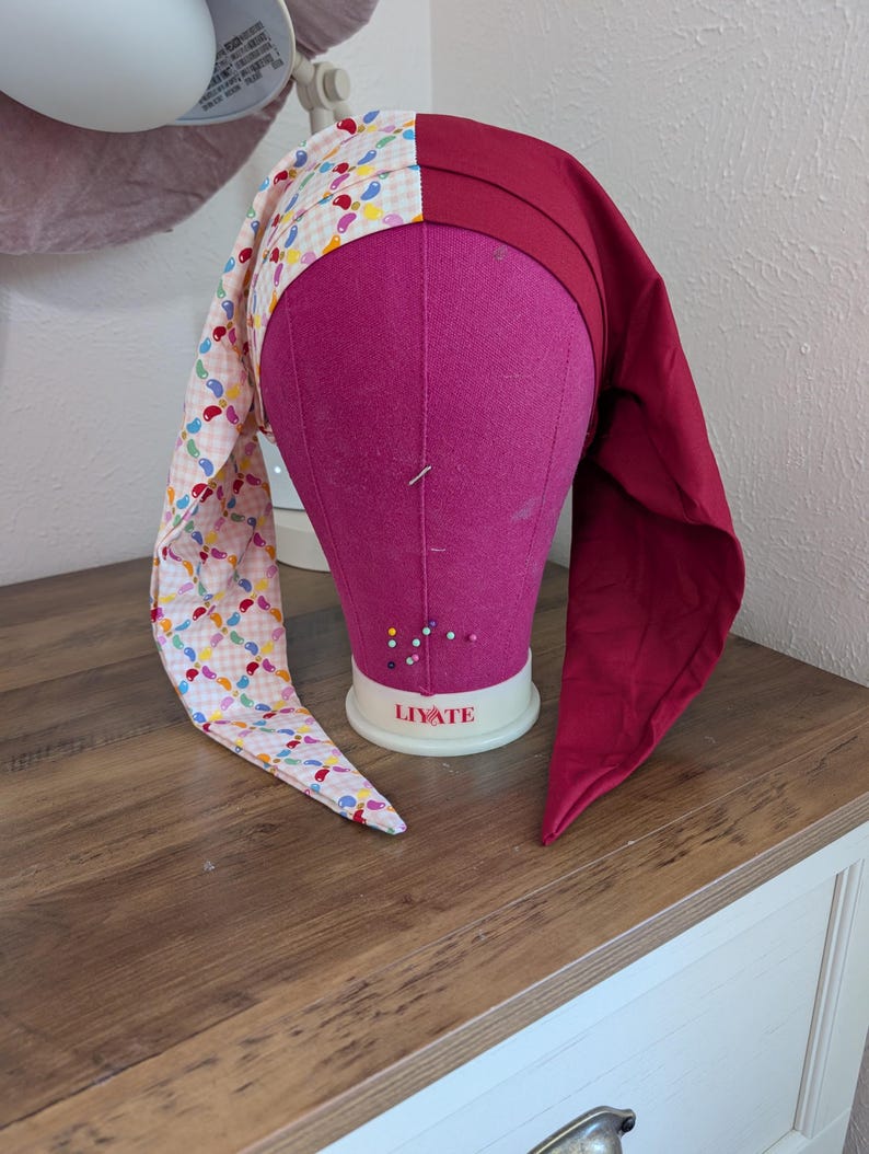 May include: A handmade jester hat with a two-tone design. One side features a patchwork of colorful teardrop shapes on a light pink background, while the other side is a solid deep red. The hat is displayed on a pink mannequin head.