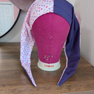 May include: A long, pointed hat with a whimsical design. The hat features a section with a pattern of small hearts in pink, blue, and red on a white background, and a solid navy blue section. The hat is displayed on a pink mannequin head.