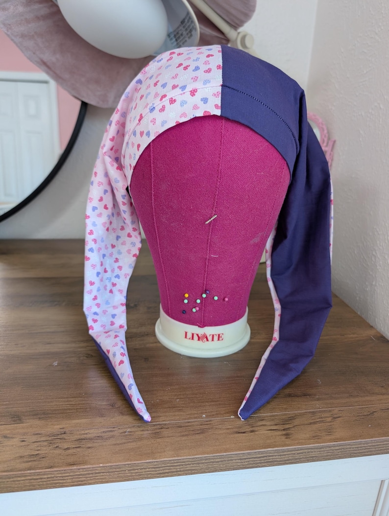 May include: A long, pointed hat with a whimsical design. The hat features a section with a pattern of small hearts in pink, blue, and red on a white background, and a solid navy blue section. The hat is displayed on a pink mannequin head.