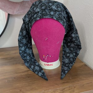 May include: A black and gray patterned head covering is draped over a pink mannequin head. The fabric features a floral design. The head covering has long, pointed ends that hang down on either side of the mannequin's face. The mannequin is on a wooden surface.