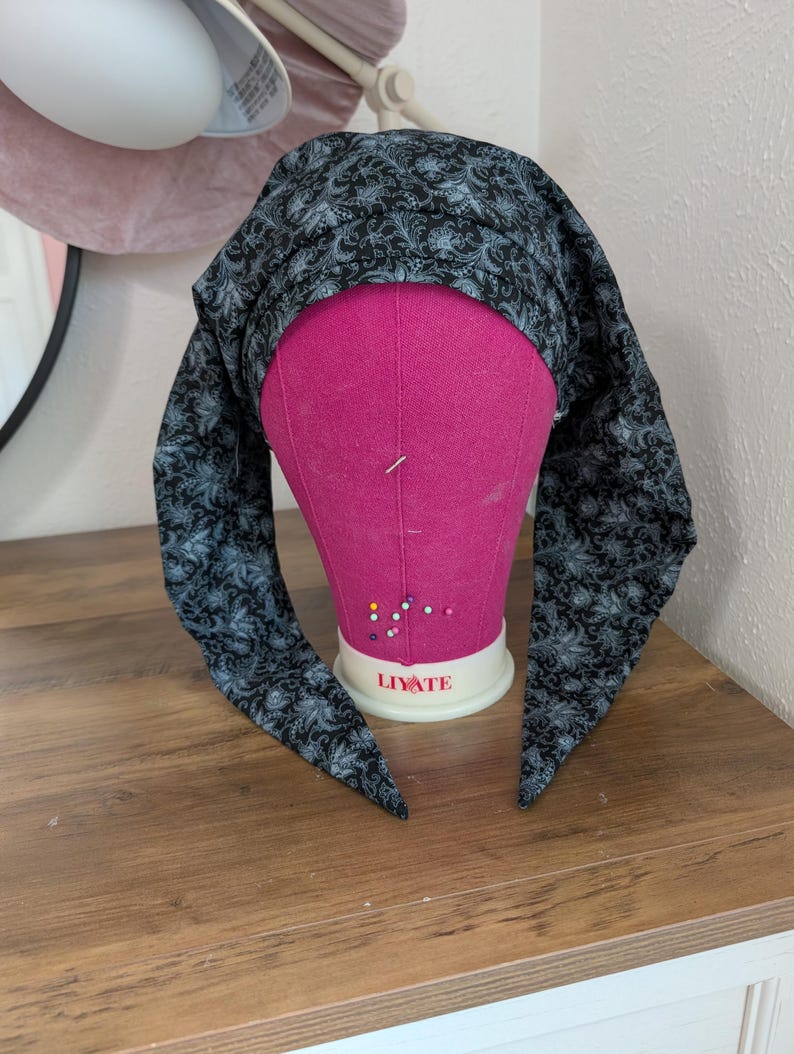 May include: A black and gray patterned head covering is draped over a pink mannequin head. The fabric features a floral design. The head covering has long, pointed ends that hang down on either side of the mannequin's face. The mannequin is on a wooden surface.
