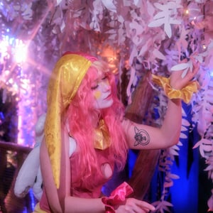 May include: A person in a pink and yellow costume with pink hair and a gold head covering. The costume includes a pink top, yellow skirt, and pink wrist cuffs. The background features white leaf decorations and a blue and purple light.