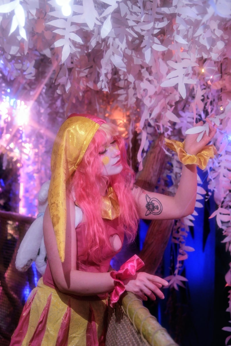 May include: A person in a pink and yellow costume with pink hair and a gold head covering. The costume includes a pink top, yellow skirt, and pink wrist cuffs. The background features white leaf decorations and a blue and purple light.