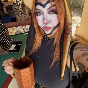 May include: A person with orange hair and theatrical makeup, wearing a black top and a gold jester hat. They are holding a wooden mug with a geometric design. The background shows a jewelry display.