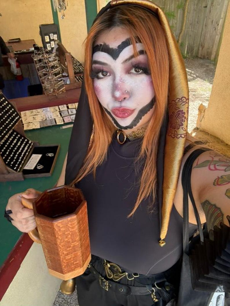 May include: A person with orange hair and theatrical makeup, wearing a black top and a gold jester hat. They are holding a wooden mug with a geometric design. The background shows a jewelry display.