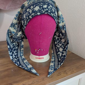 May include: A blue and cream-colored headscarf with a floral pattern is draped over a pink mannequin head. The headscarf has long, pointed ends. The mannequin head is on a wooden surface.