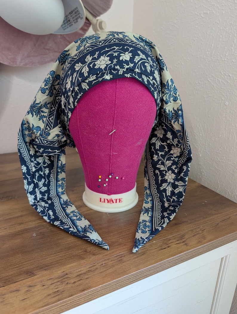 May include: A blue and cream-colored headscarf with a floral pattern is draped over a pink mannequin head. The headscarf has long, pointed ends. The mannequin head is on a wooden surface.