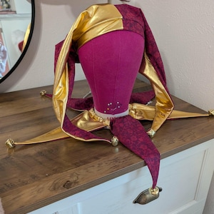 May include: A jester's hat with a gold and burgundy color scheme. The hat is on a pink mannequin head. The hat has a gold crown and burgundy fabric with a floral pattern. The hat has gold bells at the tips of the points.