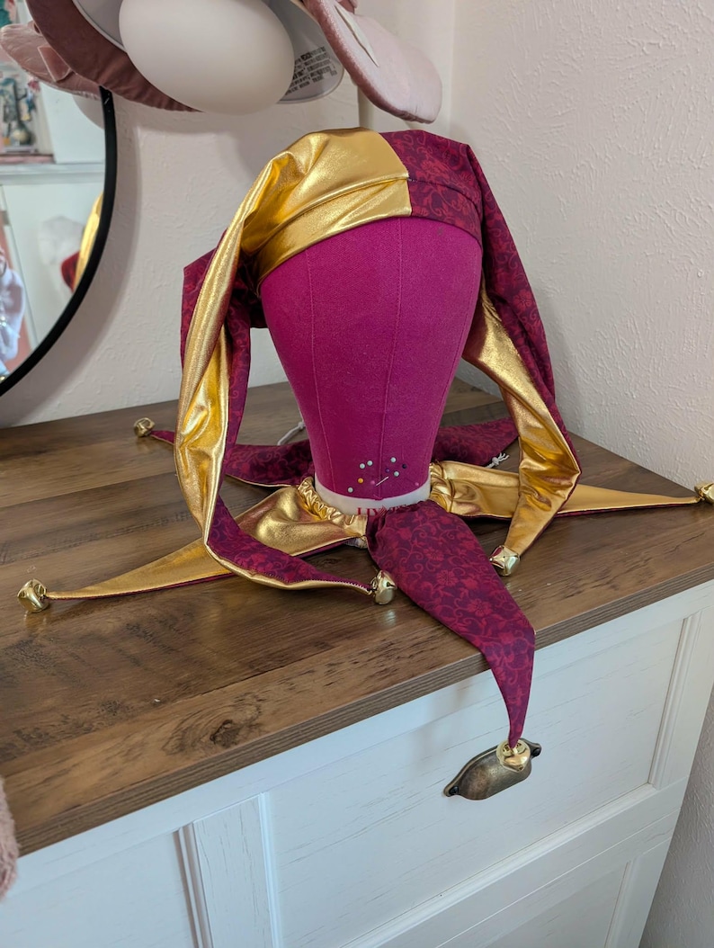May include: A jester's hat with a gold and burgundy color scheme. The hat is on a pink mannequin head. The hat has a gold crown and burgundy fabric with a floral pattern. The hat has gold bells at the tips of the points.