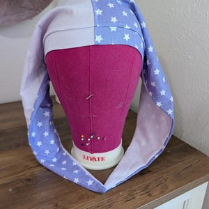 May include: A handmade sleep bonnet in a star pattern. The bonnet is two-toned, with a white and periwinkle blue fabric. The periwinkle blue fabric is decorated with white stars. The bonnet is displayed on a pink mannequin head.