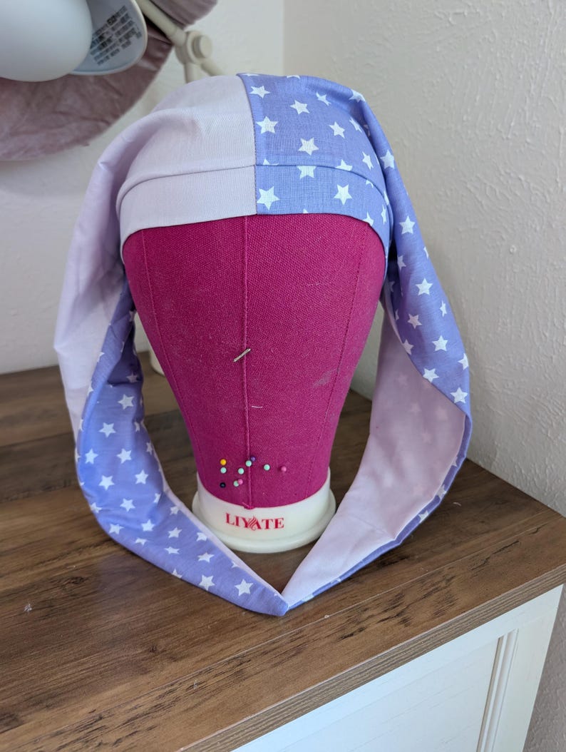 May include: A handmade sleep bonnet in a star pattern. The bonnet is two-toned, with a white and periwinkle blue fabric. The periwinkle blue fabric is decorated with white stars. The bonnet is displayed on a pink mannequin head.