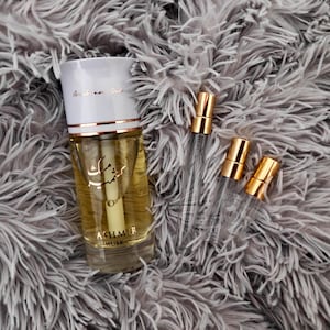 May include: A clear glass bottle with a gold cap and a white label that says "Kashmir Musk". The bottle is sitting on a fluffy grey surface. There are three small clear glass bottles with gold caps next to the larger bottle.