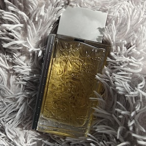 May include: A clear glass bottle of perfume with a silver cap. The bottle is filled with a golden liquid and has Arabic script on the front.