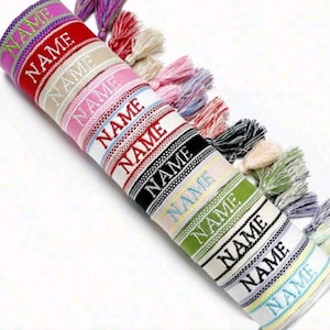Op de afbeelding: Een verzameling kleurrijke geweven armbanden met het woord "NAME" in het wit geborduurd. Elke armband heeft een ander kleurenschema, waaronder rood, roze, groen en zwart, en is versierd met bijpassende kwastjes. De armbanden zijn ideaal voor gepersonaliseerde cadeaus.