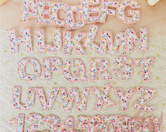 May include: A complete alphabet and number set made from fabric. The letters and numbers are white with a floral pattern in pink, yellow, and green, and are outlined with a pink border. They are displayed on a cream-colored surface.