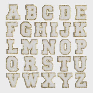 May include: A collection of white fabric letters, each outlined with gold glitter. The letters A through Z are displayed, showcasing a variety of fonts and styles. The letters are arranged in rows and columns against a plain background.