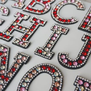 May include: Close-up of decorative rhinestone letters in various colors, including red, pink, and clear. The letters are outlined in black and are arranged on a light-colored surface. The letters are embellished with multiple rhinestones.