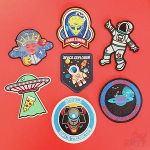 May include: A collection of embroidered space-themed patches. Designs include an astronaut, an alien, a UFO with pizza, and a space telescope. The patches feature vibrant colors and detailed stitching, perfect for decorating clothing or accessories.