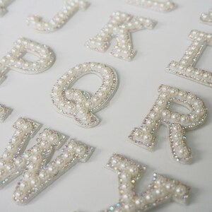 May include: Close-up of white pearl-covered letters, including the letters J, K, L, P, Q, R, V, and Y. The letters are embellished with small, iridescent rhinestones around the edges. The letters are arranged on a white surface.