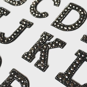 May include: Close-up of several black rhinestone letters, including J, K, and D, against a white background. The letters are embellished with small, dark rhinestones, creating a sparkling effect. The letters are likely for crafts or personalization.