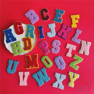 May include: A selection of colourful, embroidered fabric letter patches in various colours, including blue, red, green, and pink. The letters are arranged on a red background and a white plate.