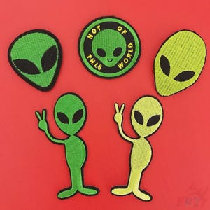 Op de afbeelding: Een verzameling geborduurde alien patches in verschillende vormen en kleuren. De patches bevatten groene alienkoppen en -figuren, waaronder een ronde patch met de tekst "NOT OF THIS WORLD".