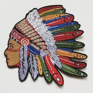 Può includere: Toppa ricamata raffigurante un copricapo nativo americano. La toppa mostra il profilo di una persona con carnagione abbronzata, adornata da un copricapo dettagliato in rosso, giallo, blu, verde e bianco. Il design include piume e cuciture intricate.