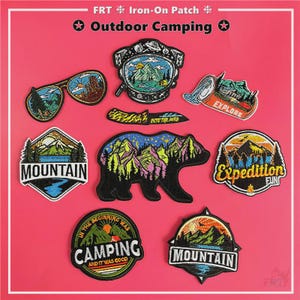 May include: A selection of iron-on patches with a camping theme. Designs include sunglasses, a knife, a bear silhouette, and mountain scenes. Text includes "Outdoor Camping", "Mountain", "Explore", and "Camping". The patches are on a pink background.