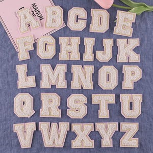 Pode incluir: Uma coleção de patches de alfabeto em estilo universitário. Cada letra é feita de um material cremoso e difuso com uma borda dourada e um verso rosa. As letras estão dispostas em uma superfície azul, com um cartão rosa com o texto "MAISON DE PELLIS".